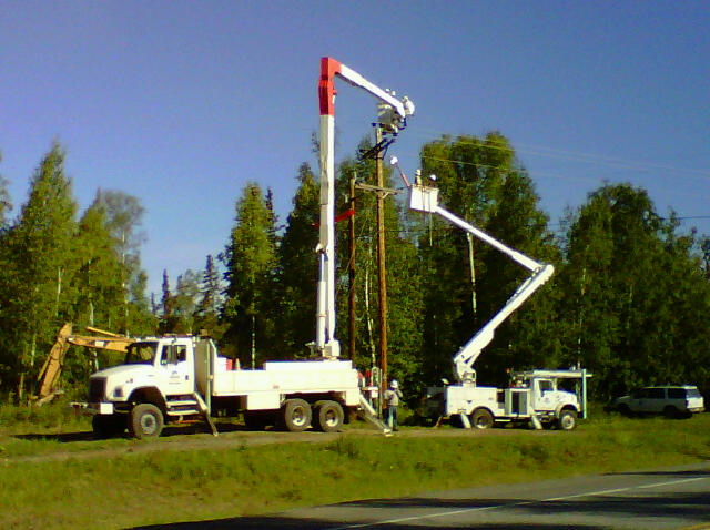 Two ALB Bucket trucks work on power lines with trees and a blue sky in the background